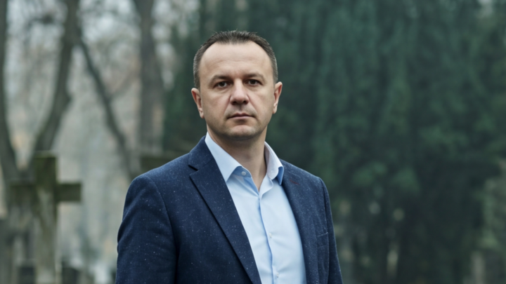 Muškarac u poslovno-ležernoj odeći stoji zamišljeno na groblju. Nosi tamni sako, svetloplavu košulju, bež chino pantalone i smeđe cipele. U pozadini se vide zamućeni nadgrobni spomenici, drveće i lišće; atmosfera deluje mirno i melanholično.